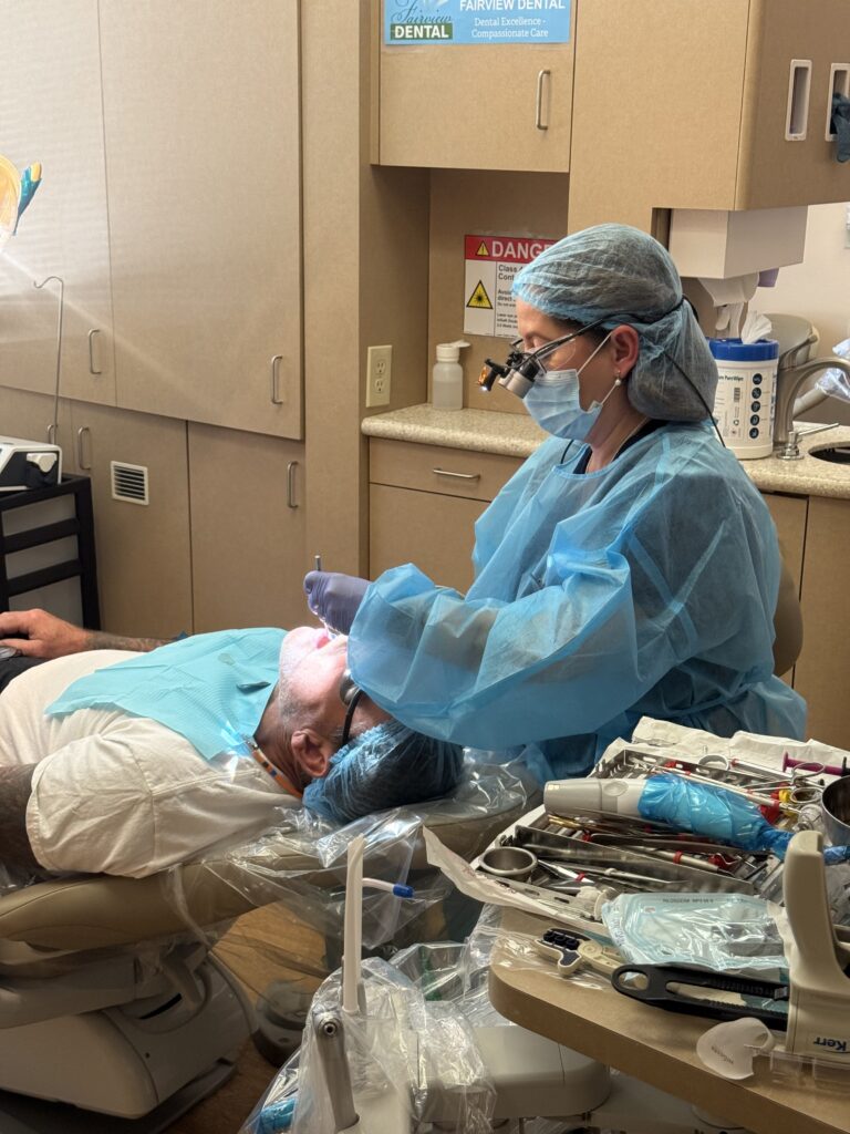 A professional dentist at Fairview Dental in Brentwood, CA, wearing full protective gear and surgical loupes, performing a precise dental procedure on a patient in a modern treatment room.
