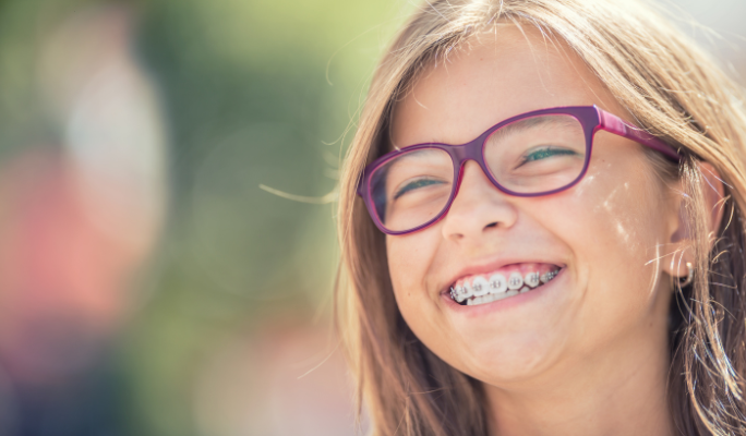 A smiling young girl with brown hair and purple glasses wearing silver metal Fastbraces to straighten her teeth.