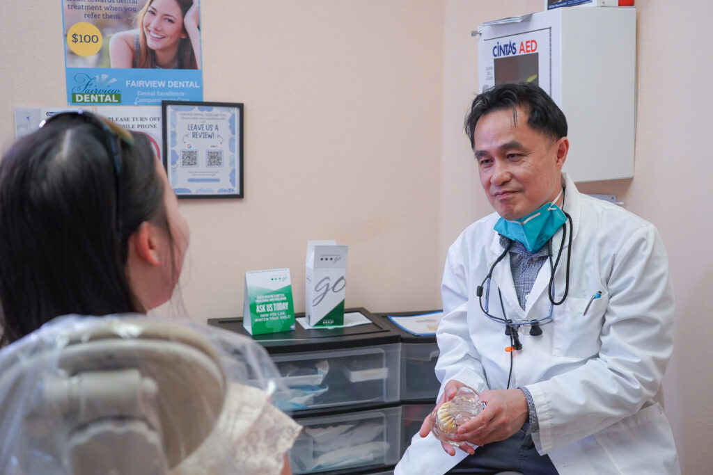 Dr. Thuc Luu of Fairview Dental Brentwood, in professional loupes and lowered mask, shows a patient a detailed anatomical model of an impacted tooth within a jawbone, explaining the extraction process during a consultation.