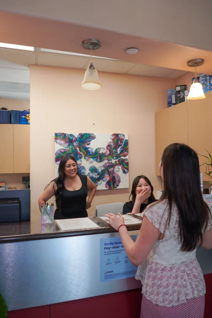 Two friendly dental reception staff members smiling and chatting warmly with a patient at the front desk counter of Fairview Dental in Brentwood, featuring a Sunbit payment plan sign.