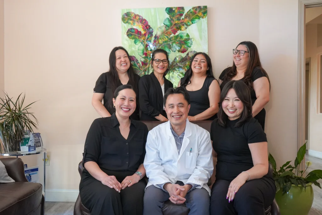 Dr. Thuc Luu sitting in a white lab coat alongside his smiling dental office staff and clinical team inside the waiting area of Fairview Dental in Brentwood.
